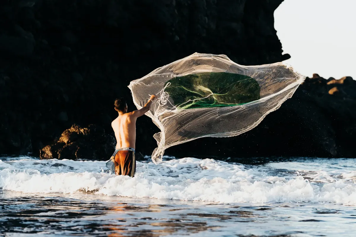 A fisherman skillfully casting a net in the shallow ocean waves by rocky cliffs, backlit by the sun.