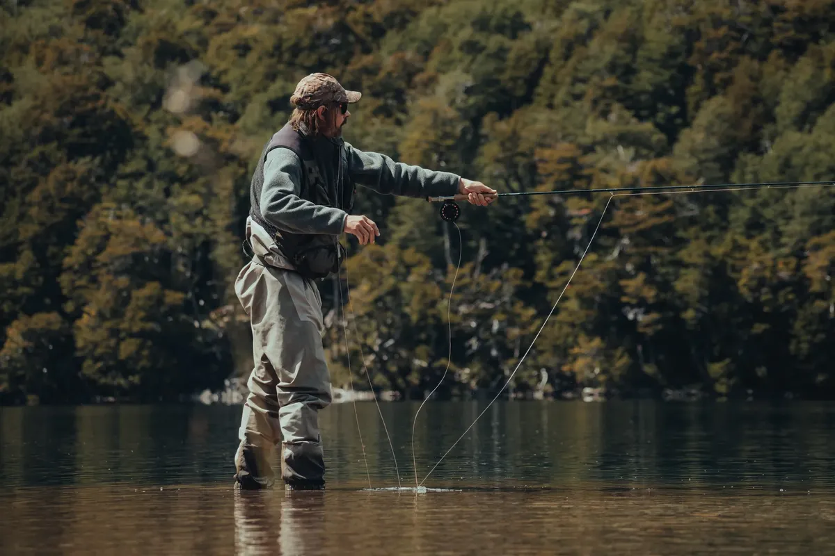 A man in waders fly fishing in a tranquil lake surrounded by lush green forest.