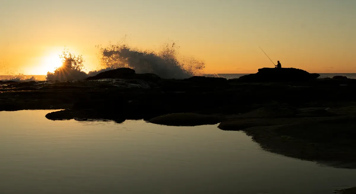 A serene sunrise silhouette of a person fishing on the rocky Sunshine Coast beach in Queensland, Australia.