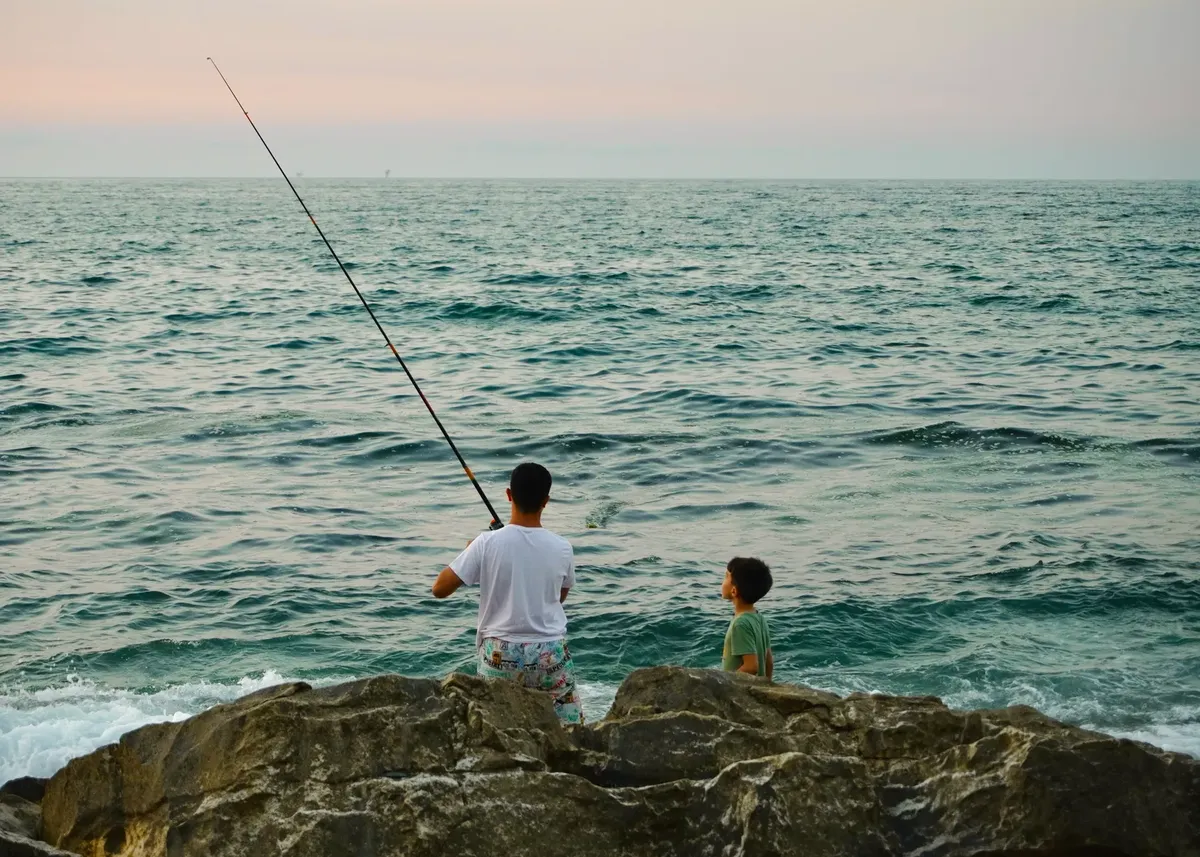 A father and son enjoy fishing on ocean rocks during a serene sunset.