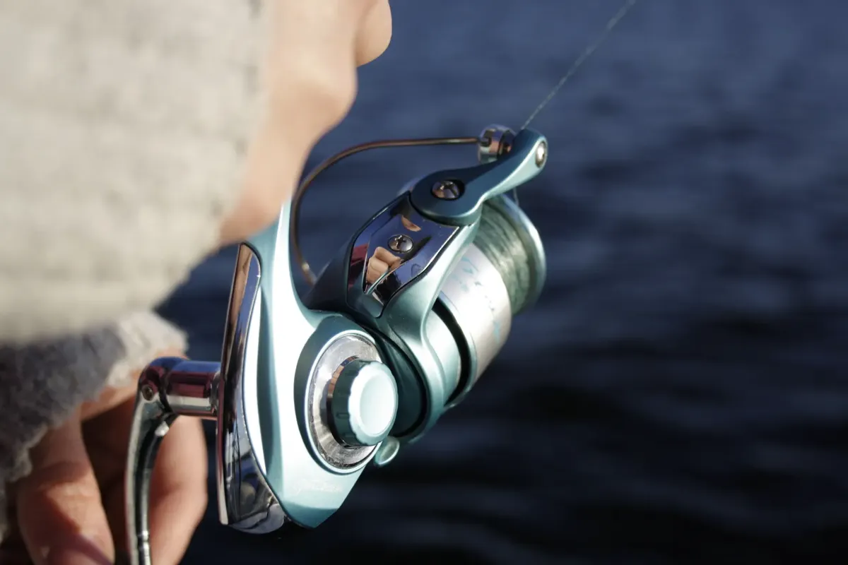 Detailed shot of a person's hand holding a fishing reel by the water, illustrating outdoor fishing activities.