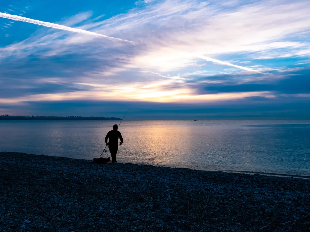 A serene scene of a silhouette fishing at sunrise by a tranquil beach.